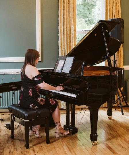Grace Coote of Reverie Music sits at a baby grand piano playing music, she is wearing a black floral dress and her back is to the camera.