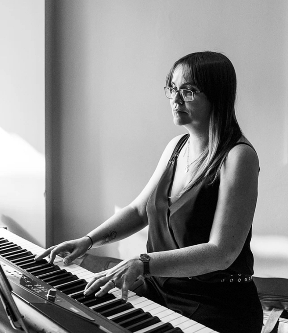 Grace Coote of Reverie Music sits at at digital piano playing music, the image is in black and white. She is facing the camera at an angle.