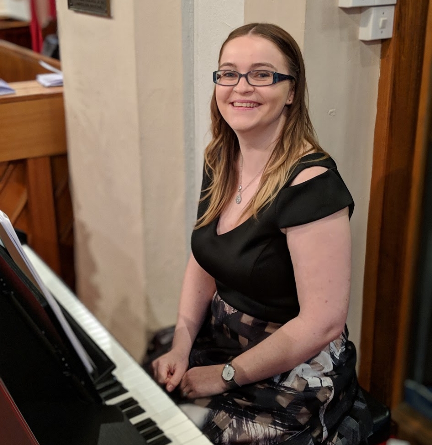 Grace Coote of Reverie Music sits a digital piano in a church, her back is to the wall and she is smiling at the camera.